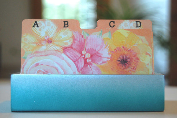 Watercolor Floral Rolodex Dividers - 2 letters per card: It is a front view of 13 dividers in a teal holder. The dividers are a coral, pink, yellow, white and green floral print.  You can see the AB and the CD dividers. There are plants in the background.