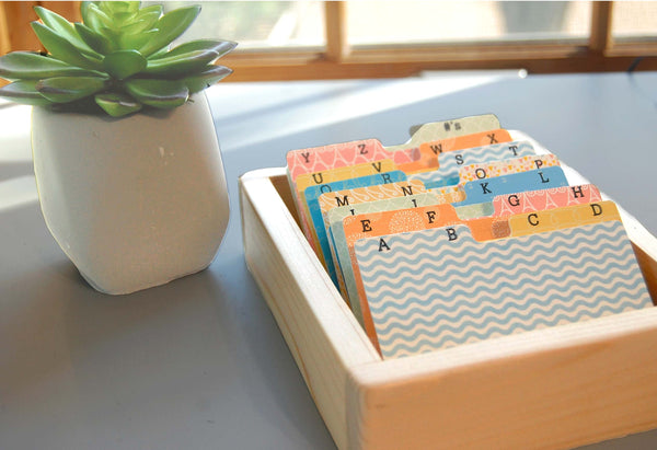 A wooden organizer tray holding colorful alphabet rolodex cards with two letters per tab, placed on a tabletop beside a small potted succulent near a window.