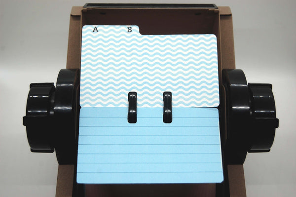A vintage Rolodex displaying a hand made Rolodex divider with AB on the tab in black capital letters on a background of wavy blue and white stripes. Beneath the divider is a lined blue Rolodex card.