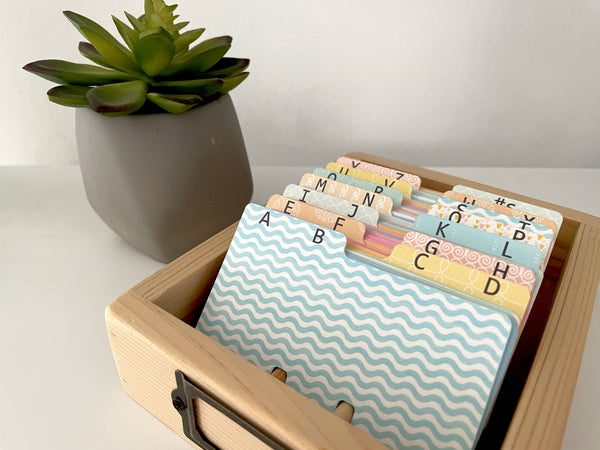 A wooden organizer box holding alphabet-indexed cards or tabs with pastel patterns, styled on a white surface beside a potted succulent.