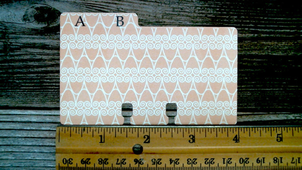 A pink and white patterned card file folder with the letter AB in black, shown on a dark wood background with a wooden ruler for scale.