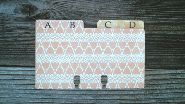 A tabbed Rolodex divider card with a pastel pink and white geometric pattern labeled A, B and behind it an orange and white patterned divider labeled C, D, displayed on a dark wood background. 