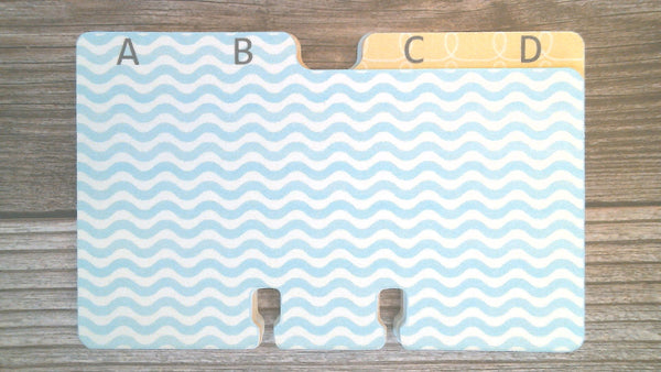 A flat organizer insert with two tabs labeled AB, and CD, featuring a light blue and white wavy pattern with a pale yellow tab at the top right, shown on a wood surface.