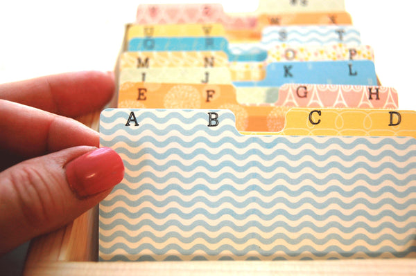 Close up of colorful Rolodex dividers in a holder. Each tab has two letters printed in black. A hand is shown holding the AB tag, which is a blue and white wavy pattern.