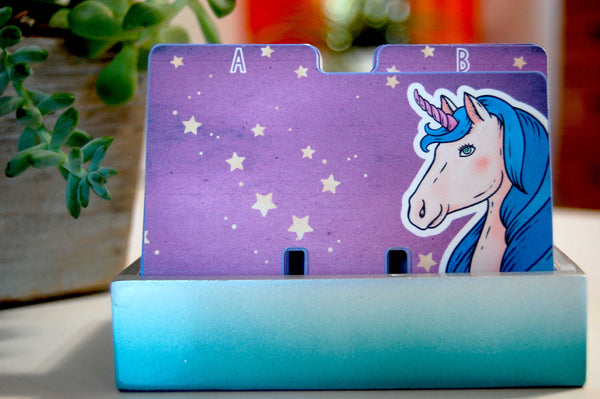A set of colorful Rolodex dividers in a teal holder with plants in the background. The Rolodex dividers are a celestial purple sky with a unicorn bust in the corner. 