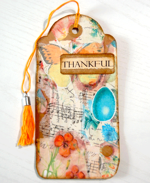 A decorated long kraft tag.  It is collaged with ripped pieces of floral paper in teal and orange.  There are musical notes, an egg sticker, and a brown tag that says "THANKFUL". There is an orange tassel through the hole in the top.