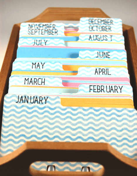 Colorful Rolodex Dividers and cards in a holder. The Rolodex dividers are a white and blue wavy stripe. They have the months of the year printed in black on the tabs. The Rolodex cards are in pastel colors of orange, green, pink and blue. 