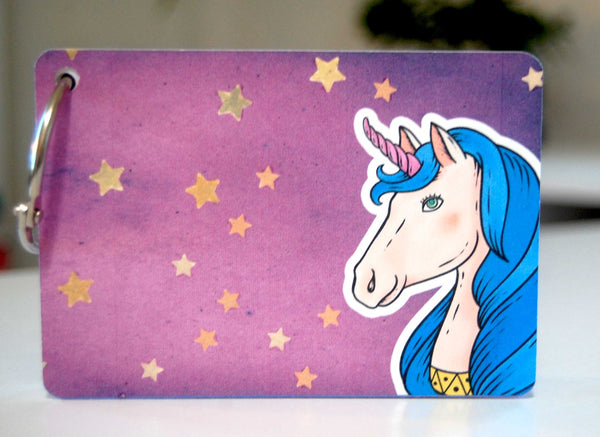 A colorful password keeper on a ring with a unicorn on the cover. the cover is a pretty purple with sparkling gold stars. The unicorn is shown from the neck up in the lower right corner. The hair is blue and the horn is pink.