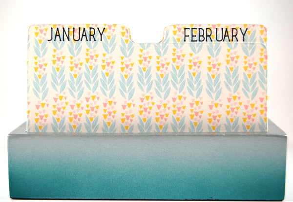Hand made Rolodex Calendar Dividers in a blue holder: The dividers are white with a delicate repeating pattern of blue leaves and tan and pink flowers. The divider on the left has "JANUARY" on the tab and the divider on the right has "February" on the tab. Both are written in black capital letters.
